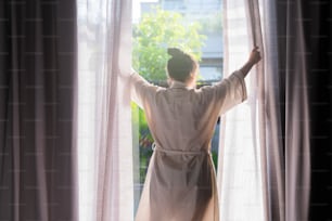 Joven mujer asiática de pie abierta cortinas blancas transparentes en la ventana, la mañana después de despertarse en el hotel dormitorio. La mujer se despierta con un fresco y abre las cortinas de las ventanas.