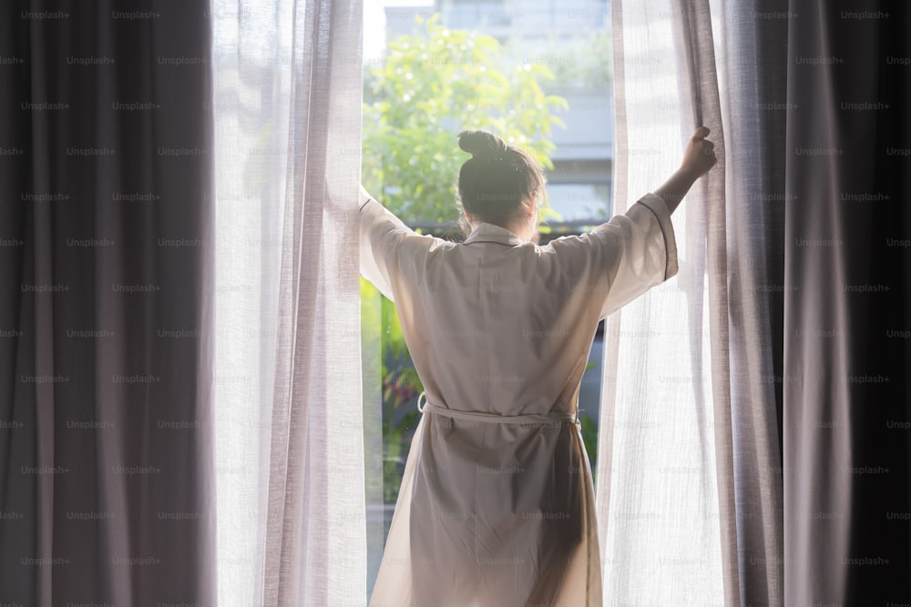 Giovane donna asiatica in piedi aperto tende bianche trasparenti alla finestra, la mattina dopo essersi svegliato nella camera da letto hotel. La donna si sveglia con un fresco e apre le tende alle finestre.
