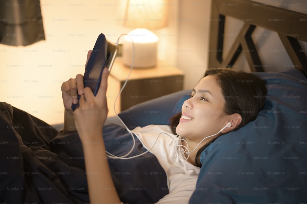 Una mujer joven está usando una tableta, viendo películas o videollamadas a sus amigos o familiares en su dormitorio, luz nocturna