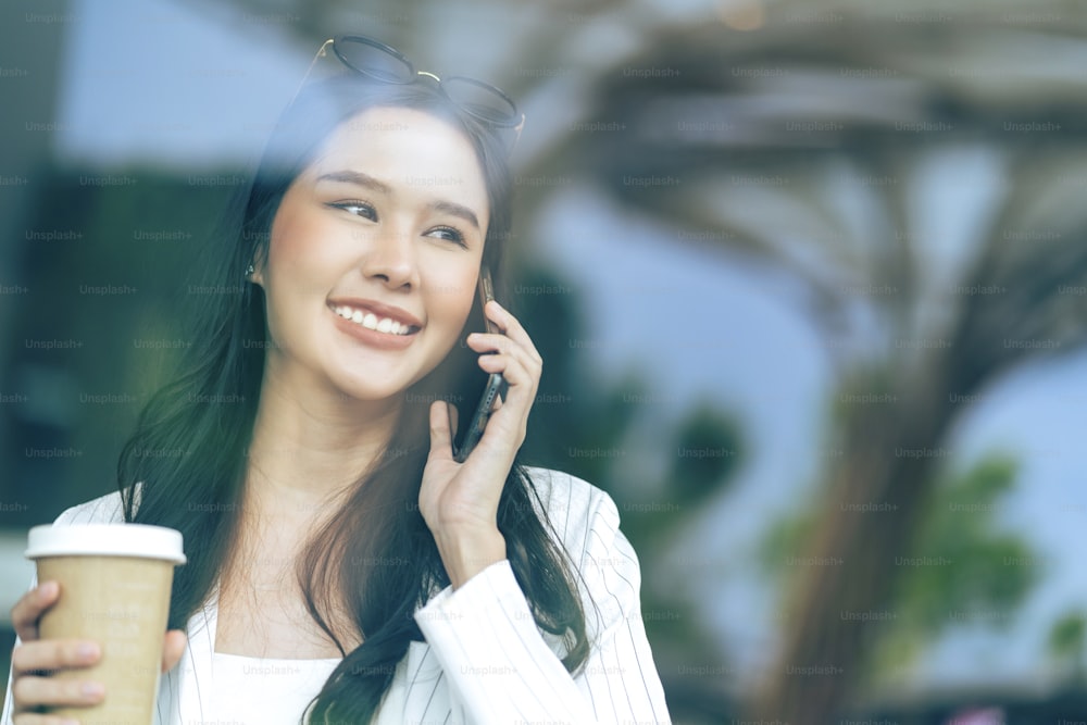 attraente asiatico femmina adulto donna d'affari formale panno felicità sorridente conversazione discutere parlare tenere la mano smartphone e tazza di caffè comunicazione con riflettere vista attraverso la finestra del caffè vetrina
