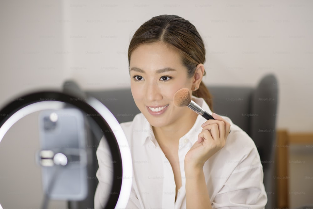 Una hermosa bloguera de maquillaje asiática está transmitiendo en vivo cómo maquillarse la cara con belleza en su hogar, concepto de belleza y tecnología.