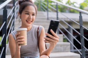 Spensierato digitale nomade asiatico femmina donna tenere la mano tazza di caffè mentre si utilizza lo smartphone chattare con il cliente remoto mentre è seduto sulla scala del parco pubblico città urbana stile di vita moderno lavoro