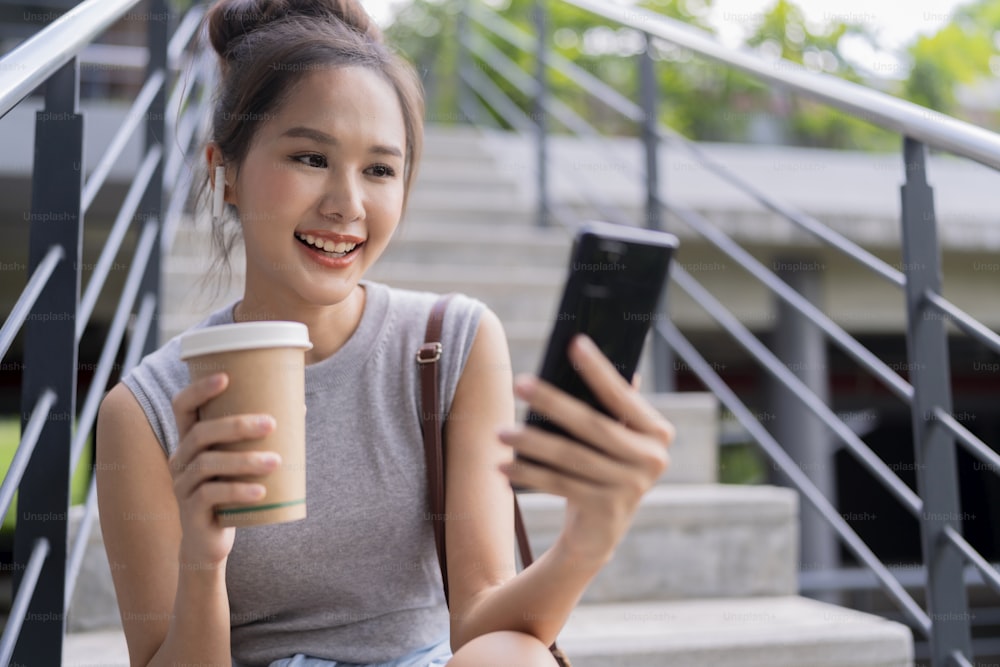carefree digital nomad asian female woman hand hold coffee cup while using smartphone chatting with remote customer while sitting on public park stair urban city modern lifestyle working