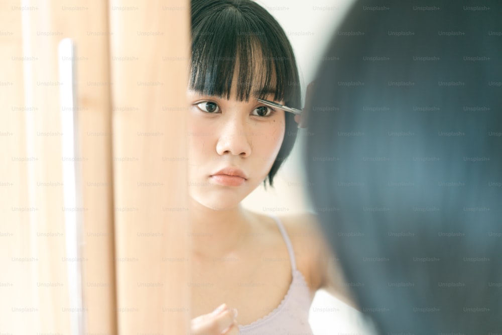 Quarantine lifestyle stay at home concept. Young adult asian woman self cutting bangs haircut with scissors. Eyes looking at a mirror. Background on day with nature light.