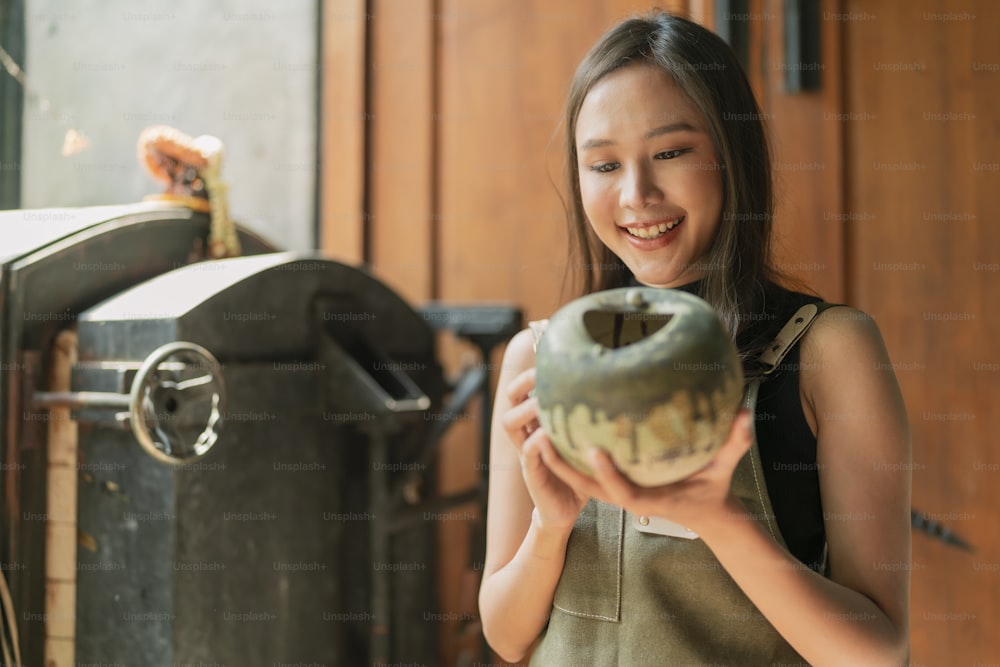 asian female adult ceramist designer hand hold ceramic clay vase handmade with cheerful pround smiling at home factory studio,asia woman wear apron looking at handmade pot with care and concentrate