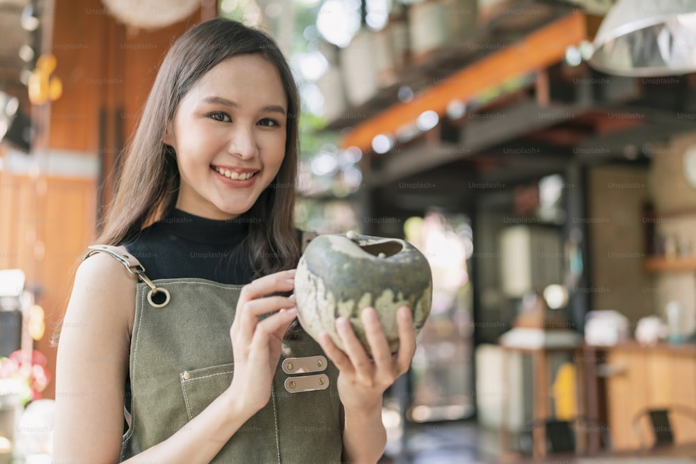 asian female adult ceramist designer hand hold ceramic clay vase handmade with cheerful pround smiling at home factory studio,asia woman wear apron looking at handmade pot with care and concentrate