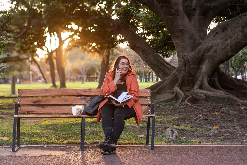 An executive plus-size woman working outdoors.  She is holding a notebook and having a telephone conversation while laughing and sitting on a bench in a public park
