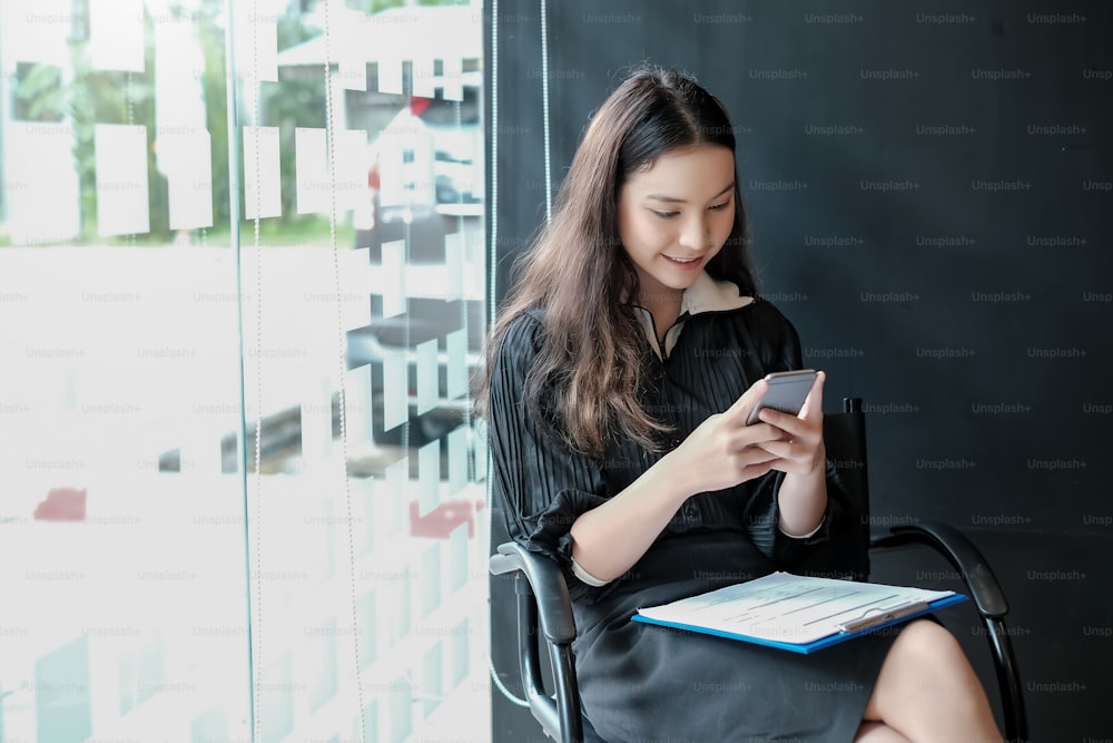 Asian woman who is sitting smiling and using a mobile phone while waiting for a job interview. Job application concept.