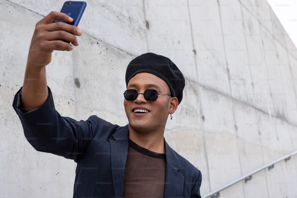 Young latin gay man with make-up on wearing a fashionable hat and sunglasses, taking selfies, isolated on a white background. LGBT. Copy space