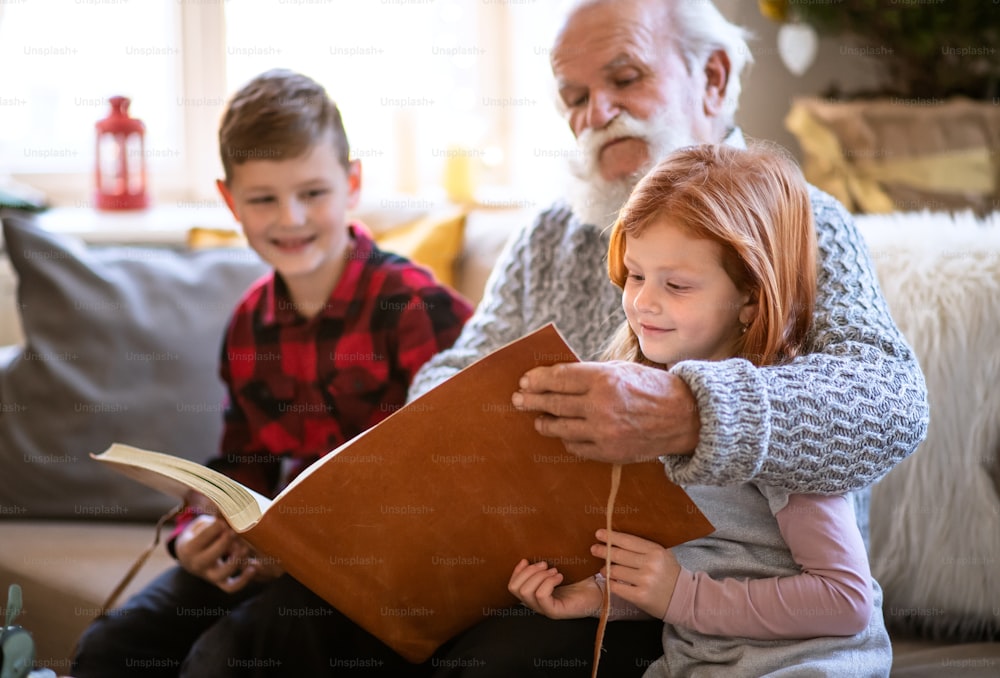 Portrait of small children with senior grandfather indoors at home at Christmas, looking at photographs.