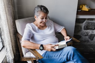 hispanic senior woman reading book and sitting on sofa at home in Mexico Latin America