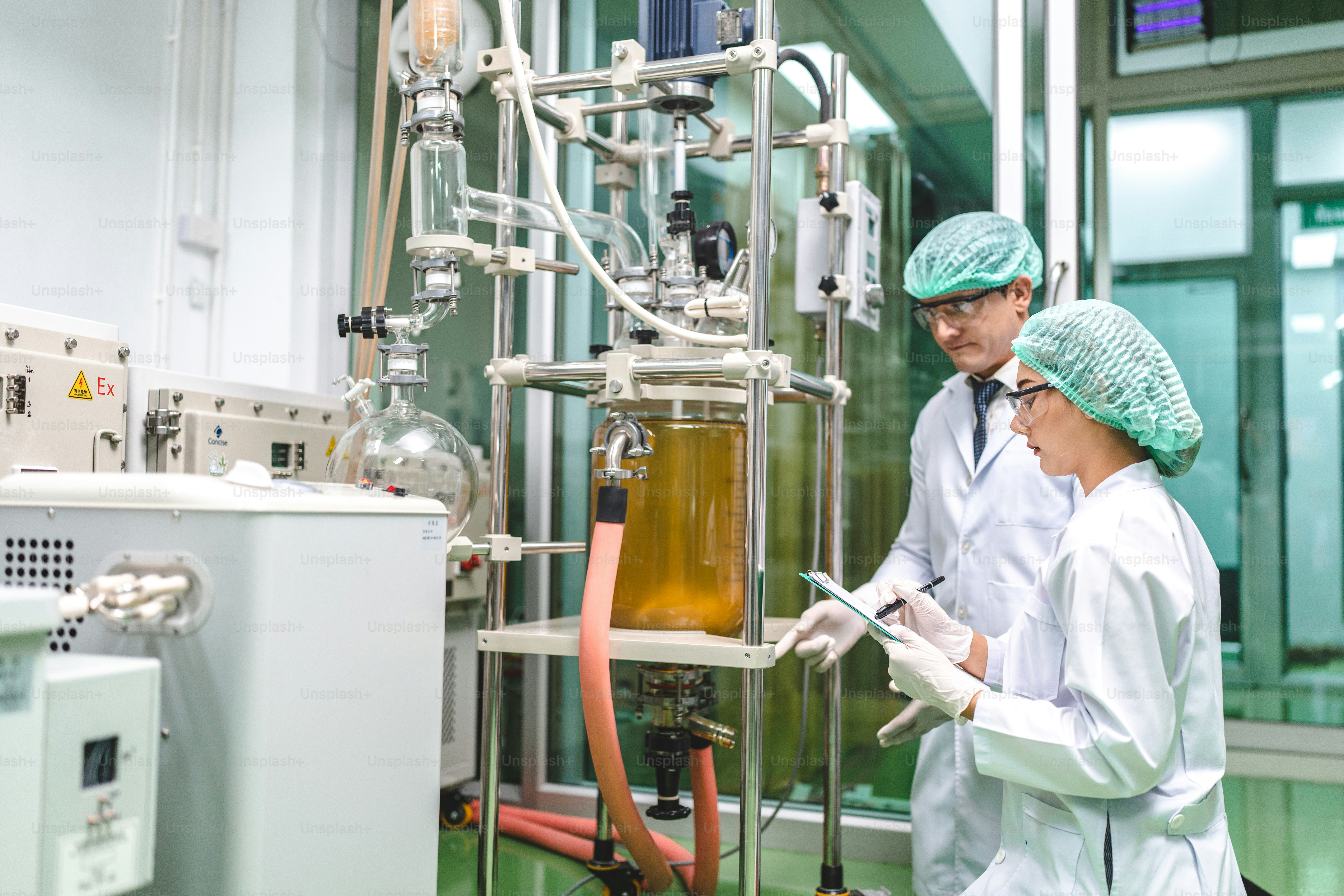Zwei Wissenschaftler in Laborkitteln und Haarnetzen überwachen einen chemischen Reaktor in einem Labor