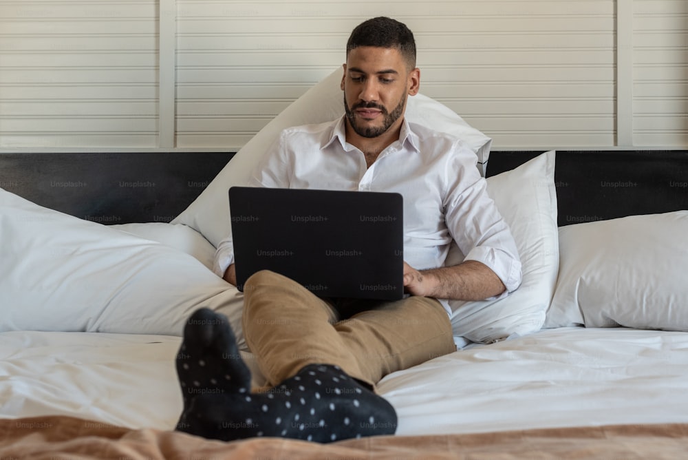 Empresario acostado en la cama trabajando en su computadora portátil en una habitación de hotel.