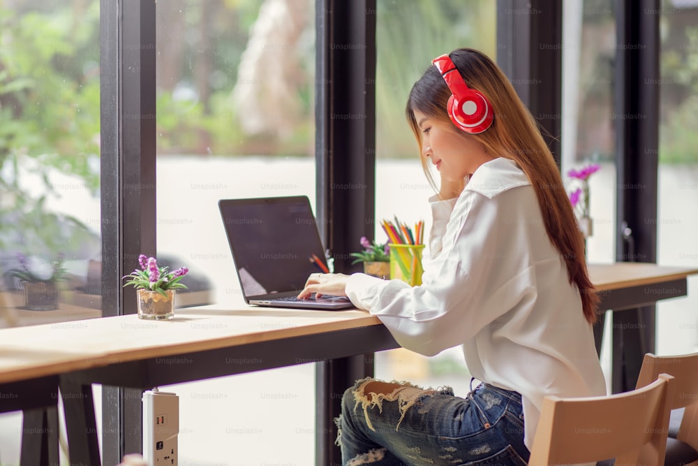 Portrait of young asian woman listening to music with headphones and using laptop computer in a cafe.