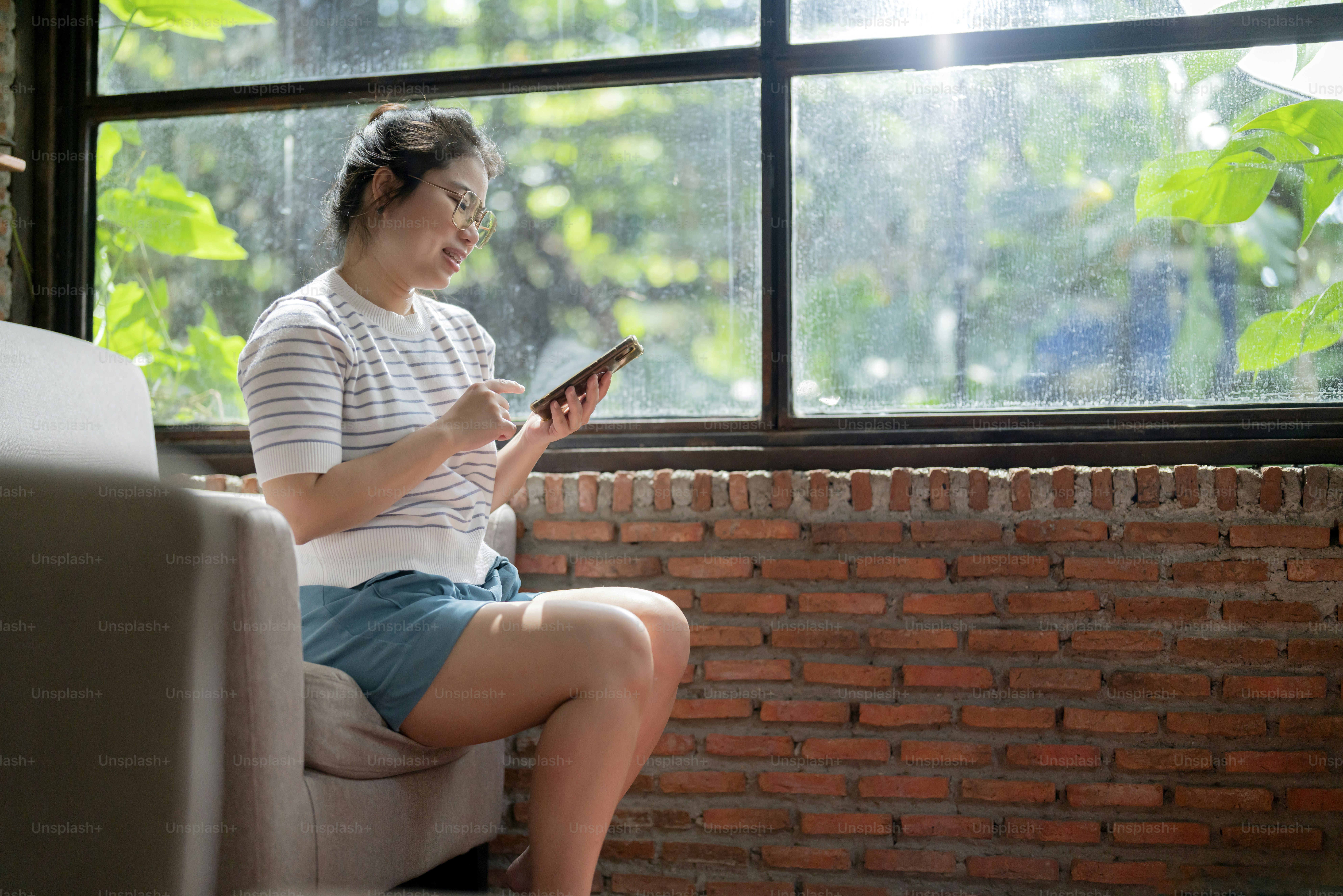 casual relax cafe lifestyle asian female woman sitting carefree peaceful emotion hand using smartphone social media browsing news or shopping online with cheerful smiling next to big window sun light