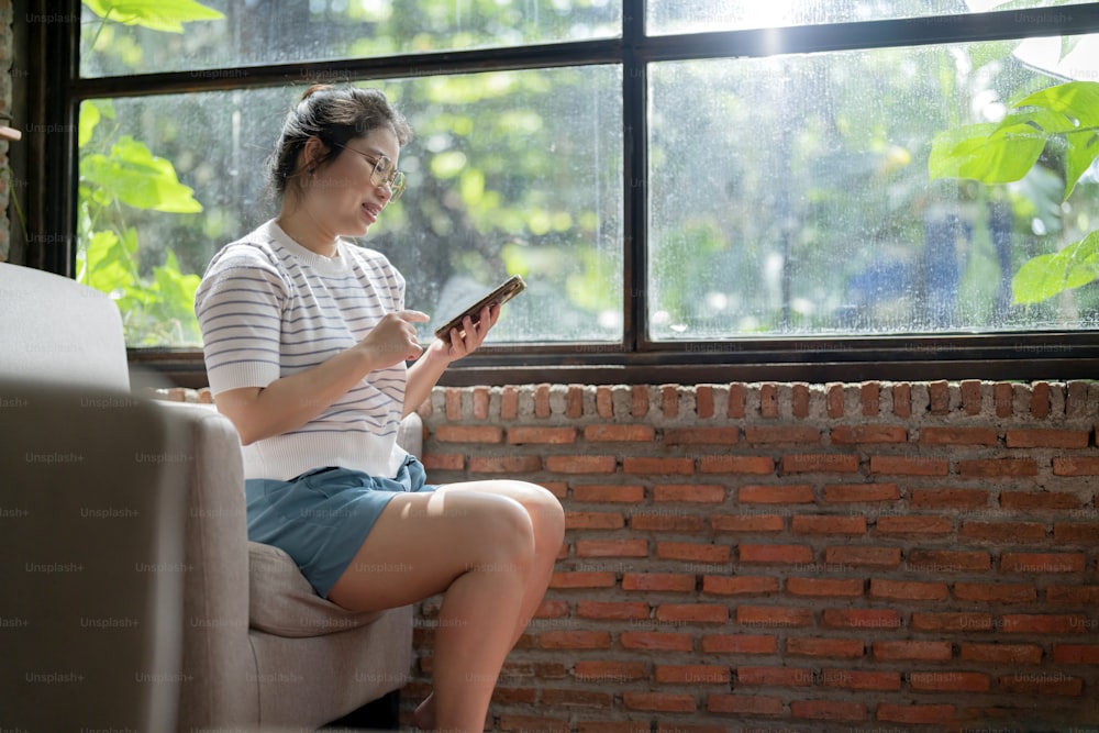 Casual Relax Café Lifestyle asiatische Frau sitzt sorglos friedliche Emotion Hand mit Smartphone Social Media Nachrichten durchsuchen oder online einkaufen mit fröhlichem Lächeln neben großem Fenster Sonnenlicht