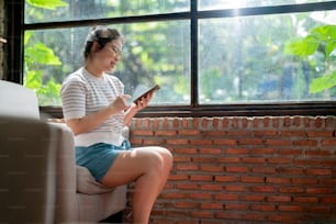 casual relax cafe lifestyle asian female woman sitting carefree peaceful emotion hand using smartphone social media browsing news or shopping online with cheerful smiling next to big window sun light