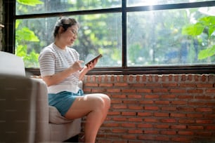 casual relax café estilo de vida mujer asiática sentada despreocupada emoción pacífica mano usando teléfono inteligente redes sociales navegando noticias o comprando en línea con alegre sonriendo junto a la luz del sol de la ventana grande