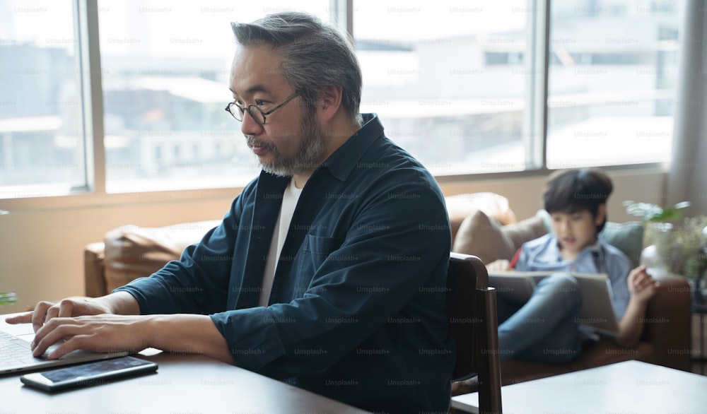 Un homme asiatique d’âge moyen utilisant un ordinateur portable travaille à domicile avec son fils dessine une image au dos.