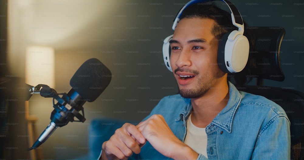Happy asia guy blogger music influencer registra un podcast sul computer con cuffie e microfono parla con il pubblico in salotto home studio di notte. Resta a casa, concetto di creatore di contenuti.