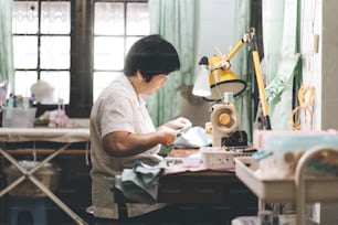 Retriement tailor southeast asian elderly crafting a cloth by sewing machine. Baby boomer generation mature working at home concept. Life moments in candid portrait authentic people.