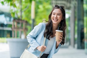 asiatico femmina casuale rilassarsi nomade digitale freelance expat programmatore smart casual panno camminando sul marciapiede città urbana con presa tazza di caffè sorridente allegro positivo sensazione downshifting stile di vita