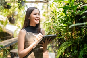 Asian female Woman gardener in apron working in a outdoor garden at home studio.Female Gardener using tablet computer for setting water drop system in greenhouse,modern greenhouse farm ideas concept