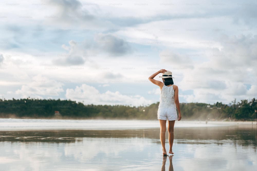 Rear view of young adult asian woman walking relax on beach sand with sunset sky. Outdoor domestic travel at andaman ocean. Phuket, Thailand.