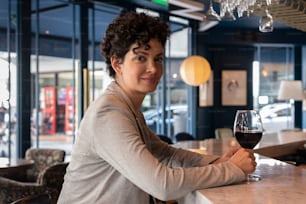 Una bella donna al bancone di un bar con un bicchiere di vino in mano che guarda la telecamera.