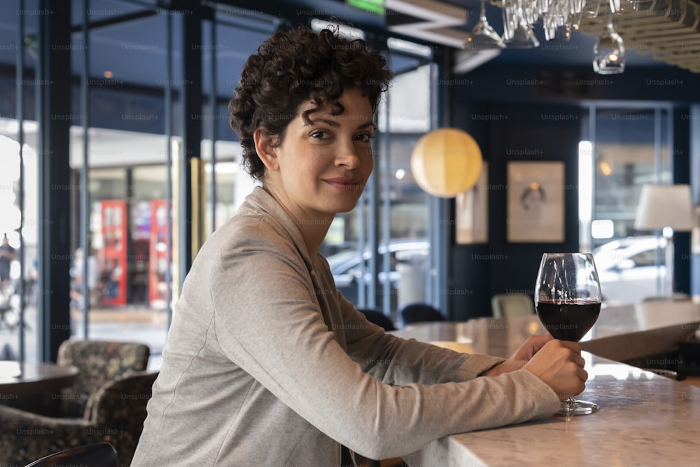 A beautiful woman at a counter of a bar with a glass of wine in her hand looking at the camera.
