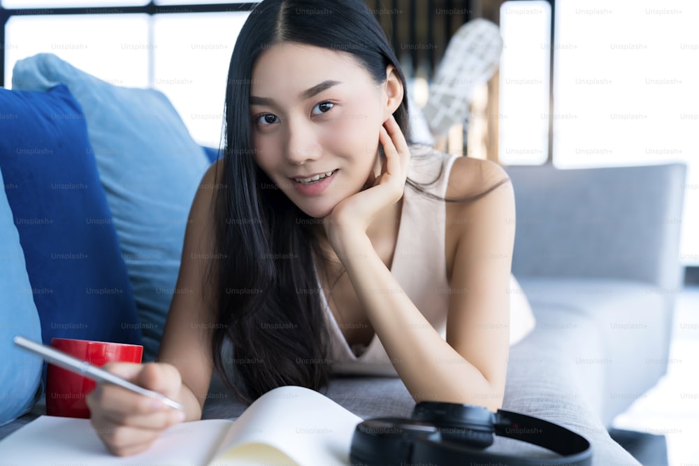 casual lifestyle asian woman work at home with notebook and coffee laydown on sofa with happiness and joyful