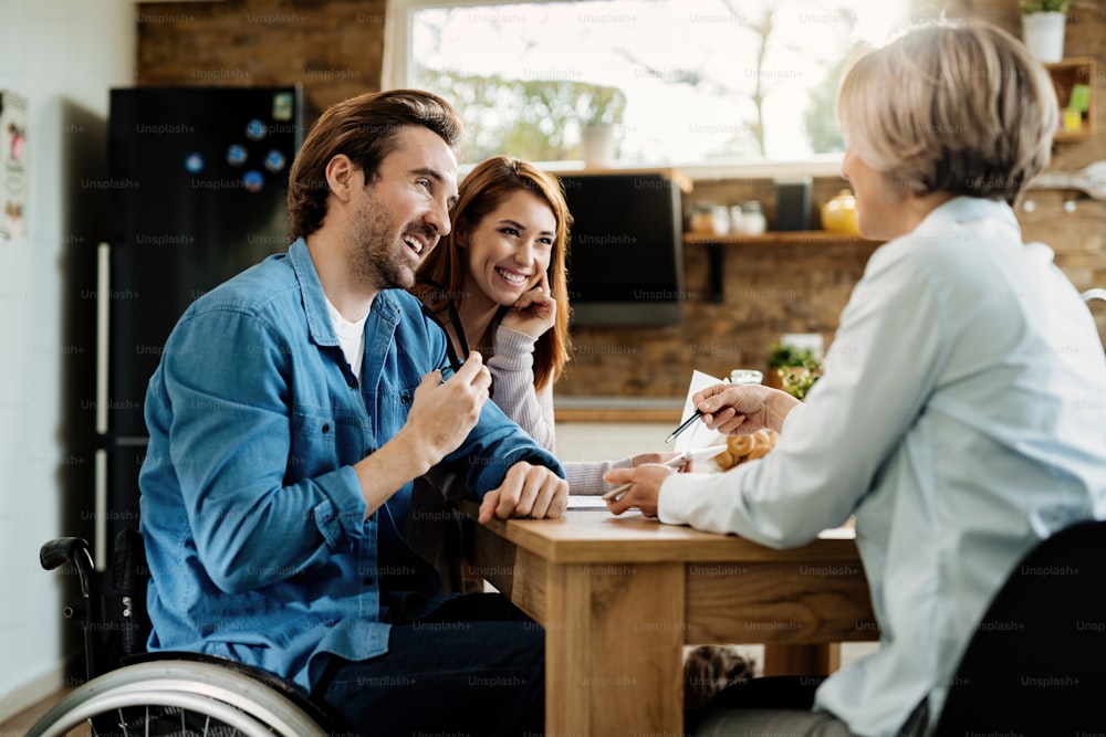 Hombre feliz en silla de ruedas y su esposa usando una tableta digital con su asesor financiero durante una reunión en casa.