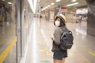 Uma jovem e bonita asiática está usando máscara de proteção em pé na estação de metrô, Nova viagem normal, proteção contra a covid-19, viagens seguras, viagens sob COVID-19, conceito de viagem pós-pandemia.