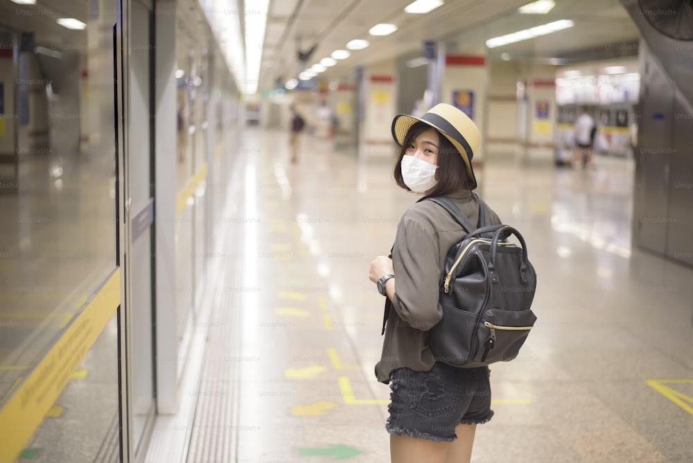 젊고 예쁜 아시아 여성이 지하철역, 뉴노멀 여행, 코로나19 보호, 안전한 여행, COVID-19 하의 여행, 팬데믹 이후 여행 개념에 서 있는 보호 마스크를 쓰고 있다.