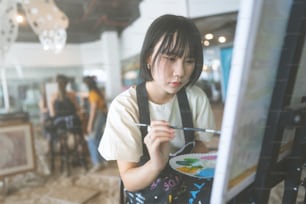 Glückliche junge asiatische Frau, die im Workshop Kunstunterricht Pinsel auf Leinwand malt. Menschen Freizeit mit Kreativität Bildung Lebensstil für psychische Gesundheit Konzept.