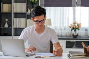 Un joven asiático está tomando notas en la mesa de la computadora portátil en casa.