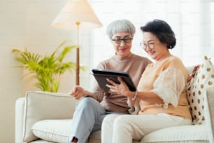 visit house of two senior old mature female friends looking through old photos digital album together with tablet device on sofa in living room at home old friend Spending Quality Time Together