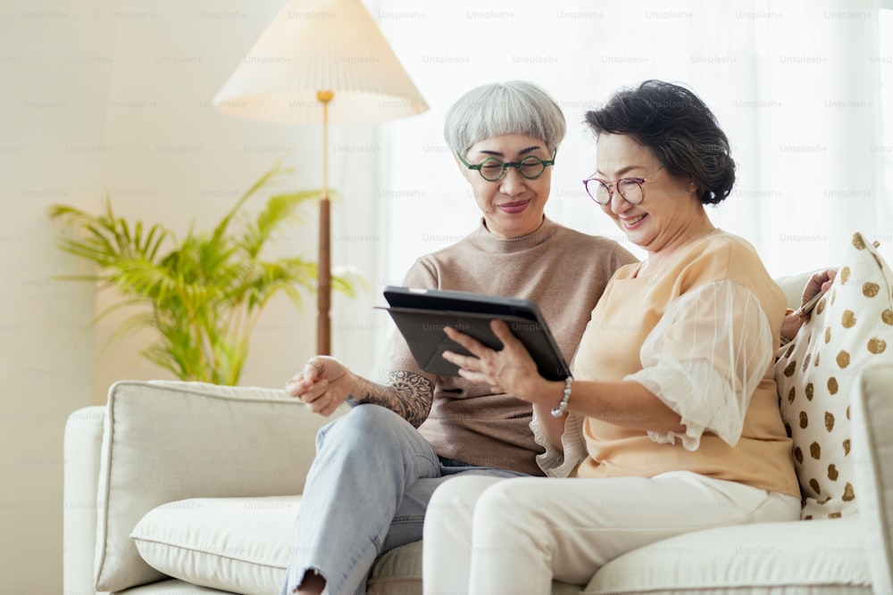 visit house of two senior old mature female friends looking through old photos digital album together with tablet device on sofa in living room at home old friend Spending Quality Time Together