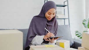 Jovem empresária muçulmana da Ásia verificar ordem de compra de produtos em estoque e economizar para o trabalho do computador tablet em home office. Pequeno empresário, entrega de mercado on-line, conceito freelance de estilo de vida.