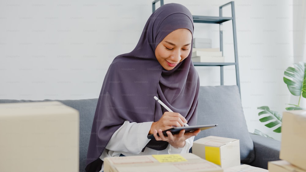 若いアジアのイスラム教徒のビジネスウーマンは、在庫のある製品の発注書を確認し、ホームオフィスでタブレットコンピュータに保存します。中小企業の経営者、オンライン市場への配送、ライフスタイルのフリーランスのコンセプト。