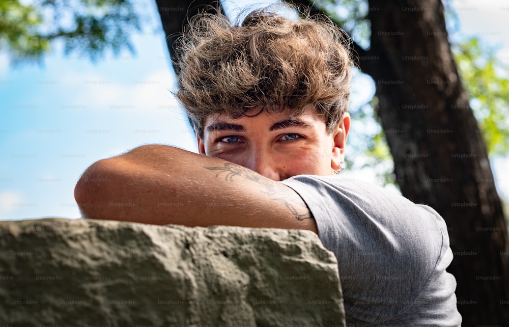 Young man falls his head down on his arms, smiling and looking at the camera in a public park