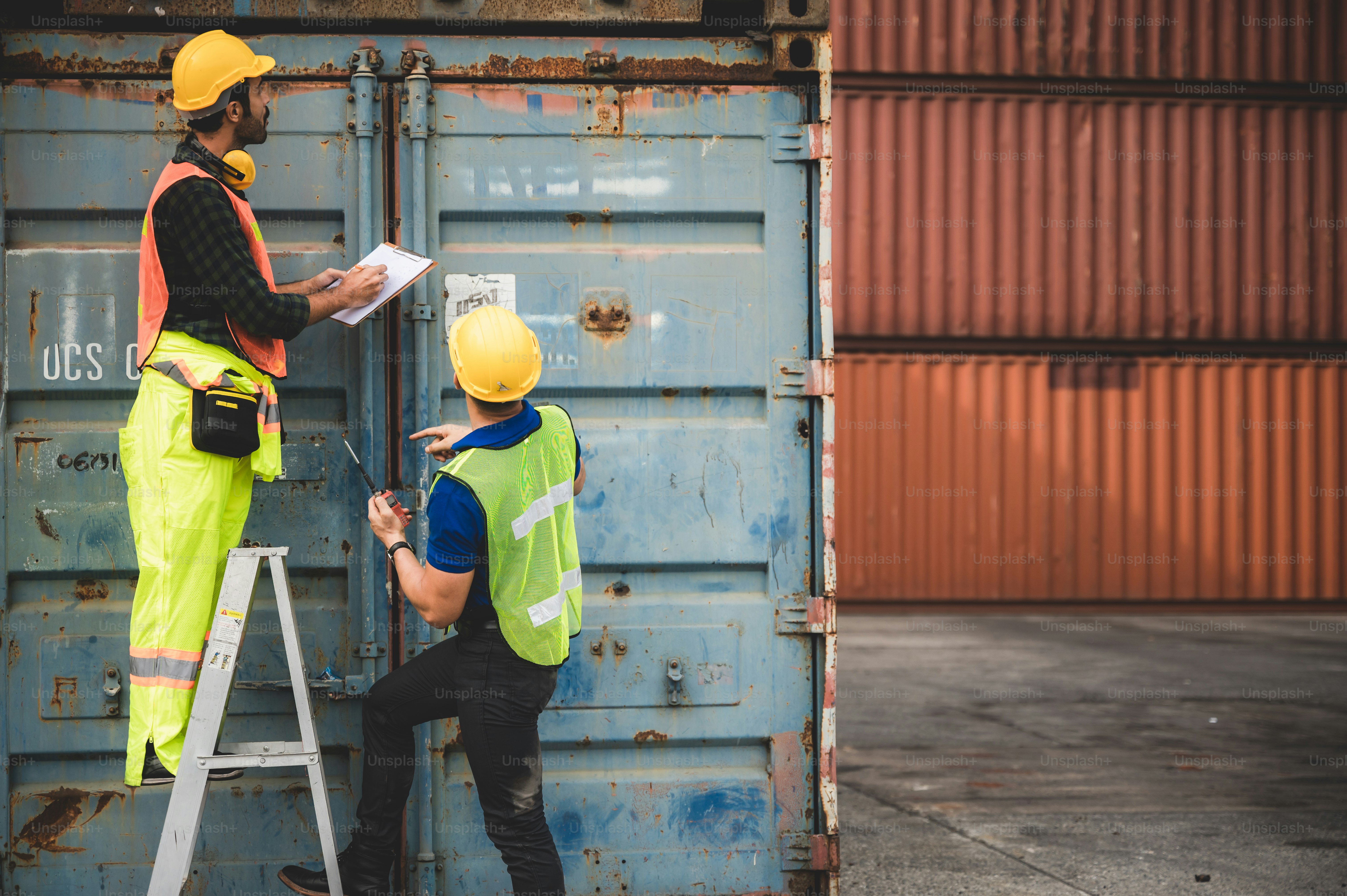 Dos trabajadores con cascos y chalecos de seguridad inspeccionando un contenedor de transporte azul