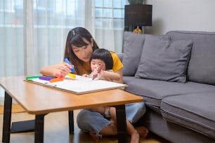 A young mom helping daughter drawing with colored pencils in living room at home.
