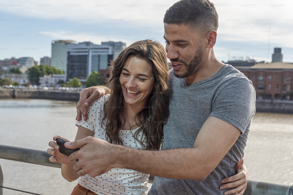 Happy young couple holding a modern smartphone in hands. They are by the river in a beautiful city.