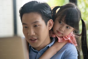 Père asiatique en chemise bleue travaillant à domicile avec sa petite fille mignonne. Un enfant asiatique étreint son père par derrière pendant que son père travaille sur un ordinateur portable.