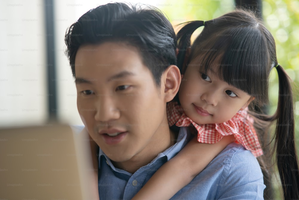 Asian father on blue shirt working from home with his little cute girl daughter. Asian kid hug his dad from behind while her father is working on a laptop.