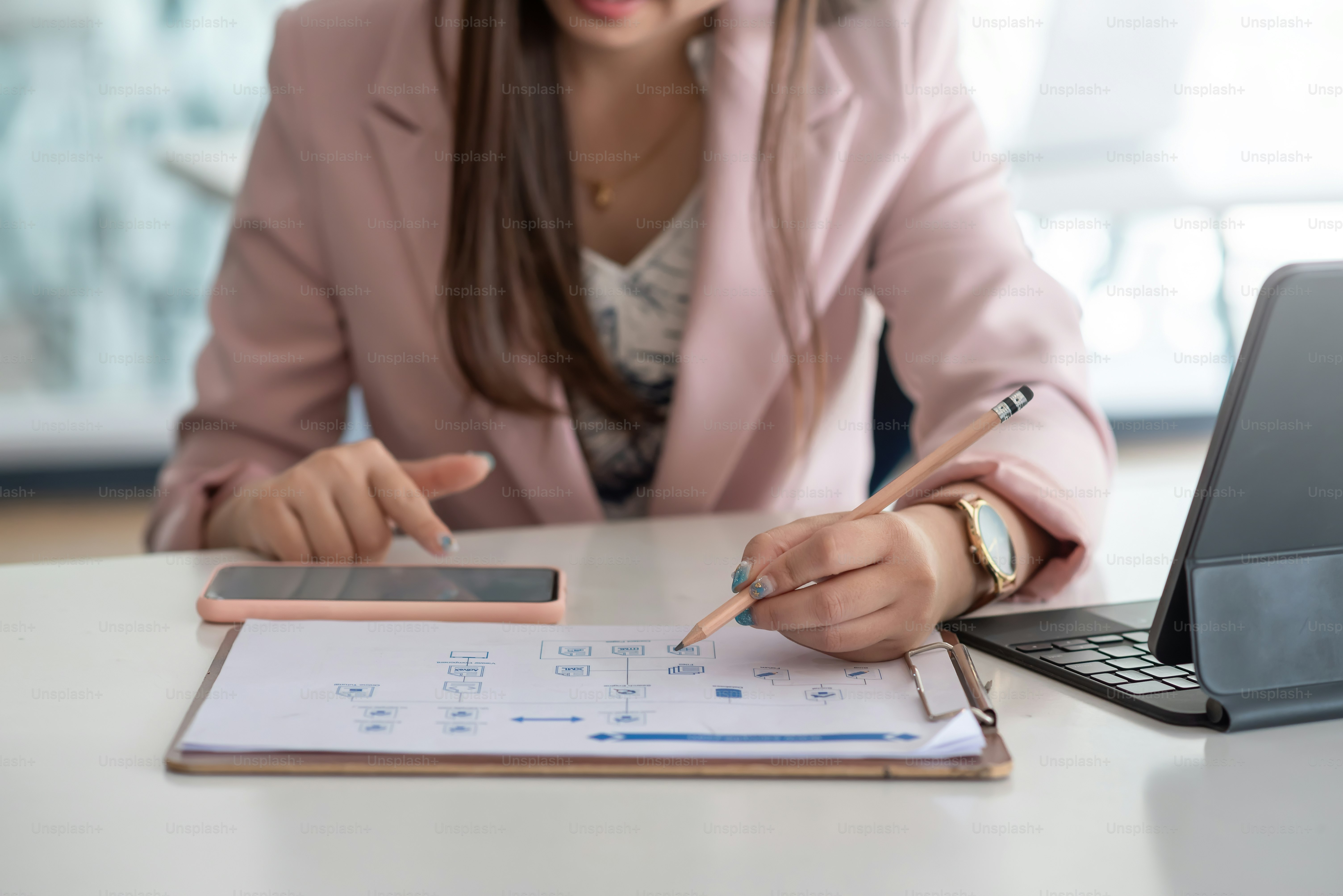 Close-up of a businesswoman holding a pencil pointing at a document with a smartphone at an office desk.
