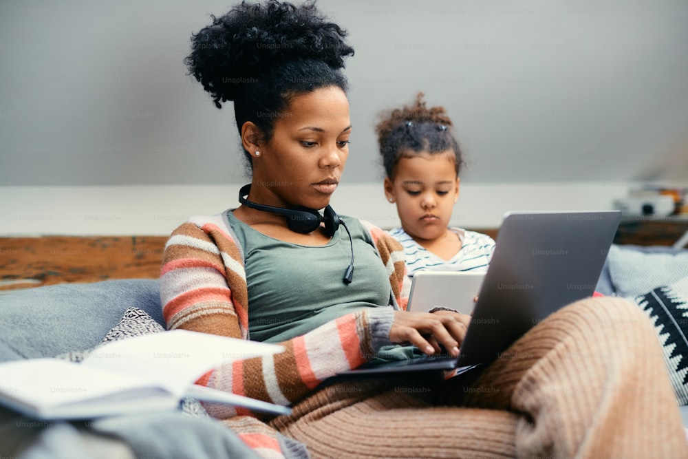 Afroamerikanisches kleines Mädchen mit Touchpad, während es neben ihrer Mutter sitzt, die zu Hause am Laptop arbeitet. Der Fokus liegt auf der Mutter.