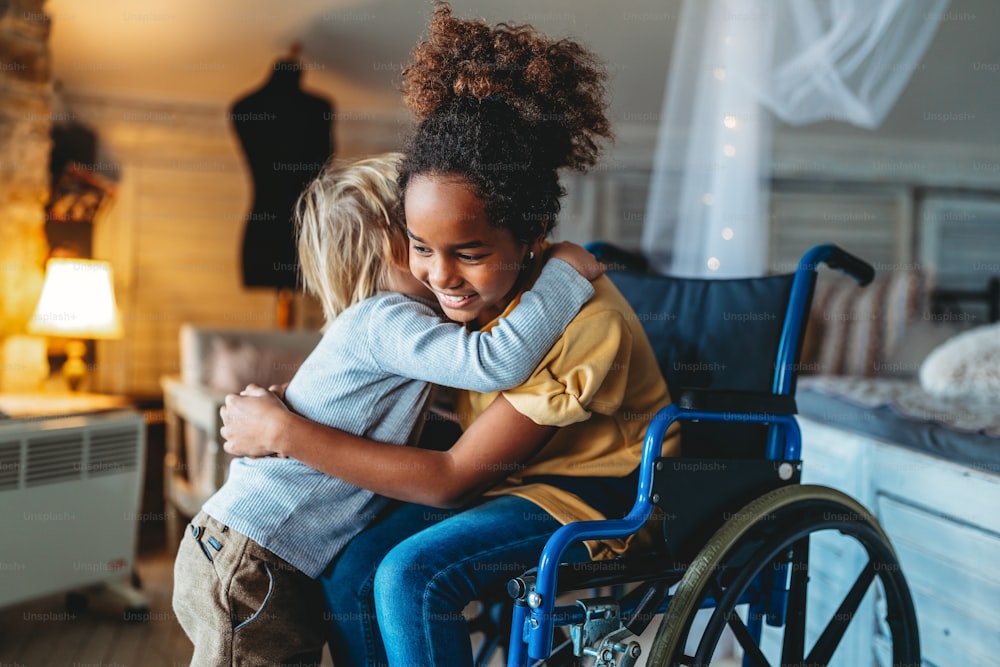 Famille aimante multiethnique heureuse. Petite fille souriante handicapée en fauteuil roulant étreignant son petit frère à la maison