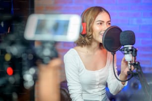 Una giovane cantante sorridente che indossa cuffie con un microfono mentre registra una canzone in uno studio musicale con luci colorate.